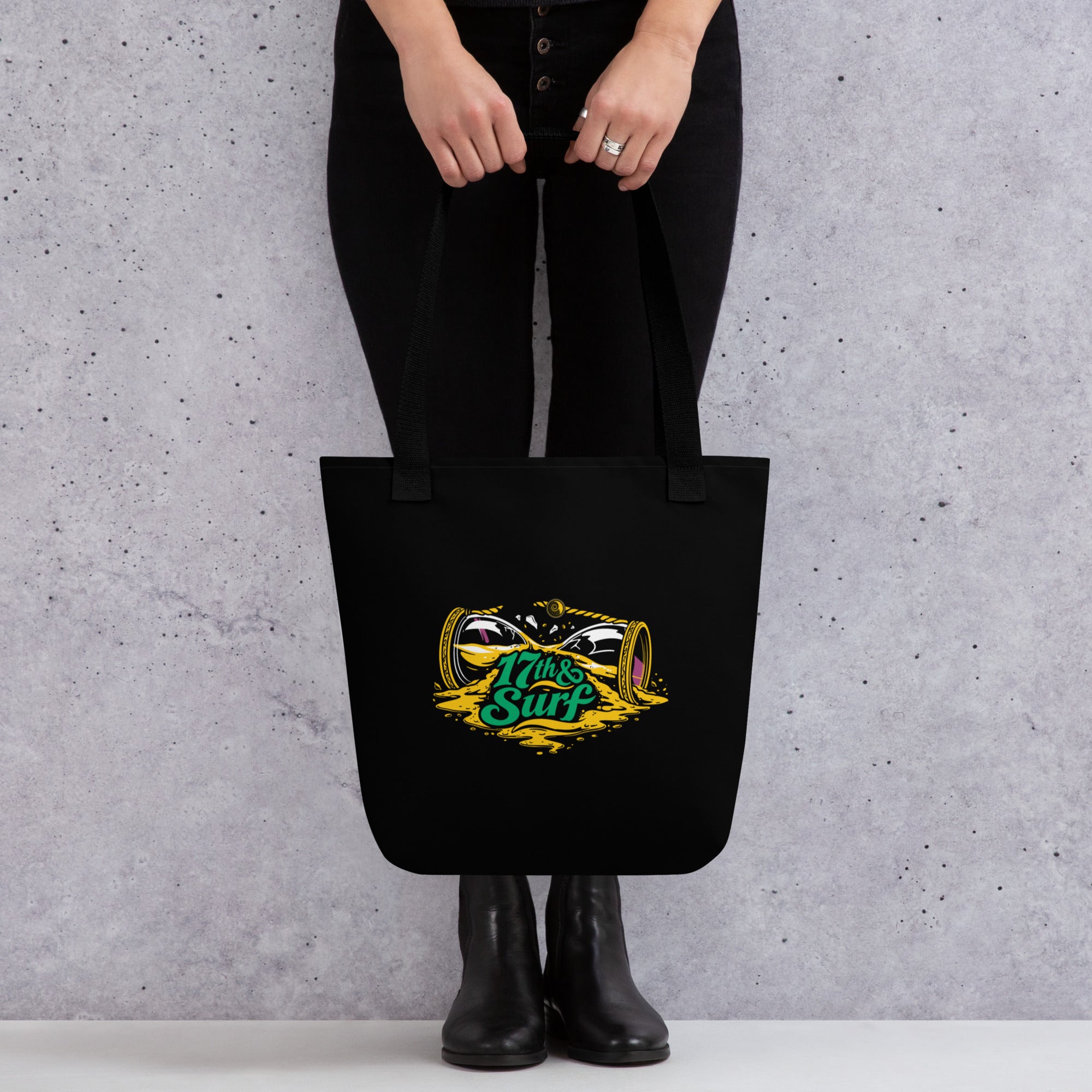 "Take Time To Decompress" black tote bag, close-up of the back panel featuring a gold and teal "17th & Surf" hourglass logo – 17th & Surf