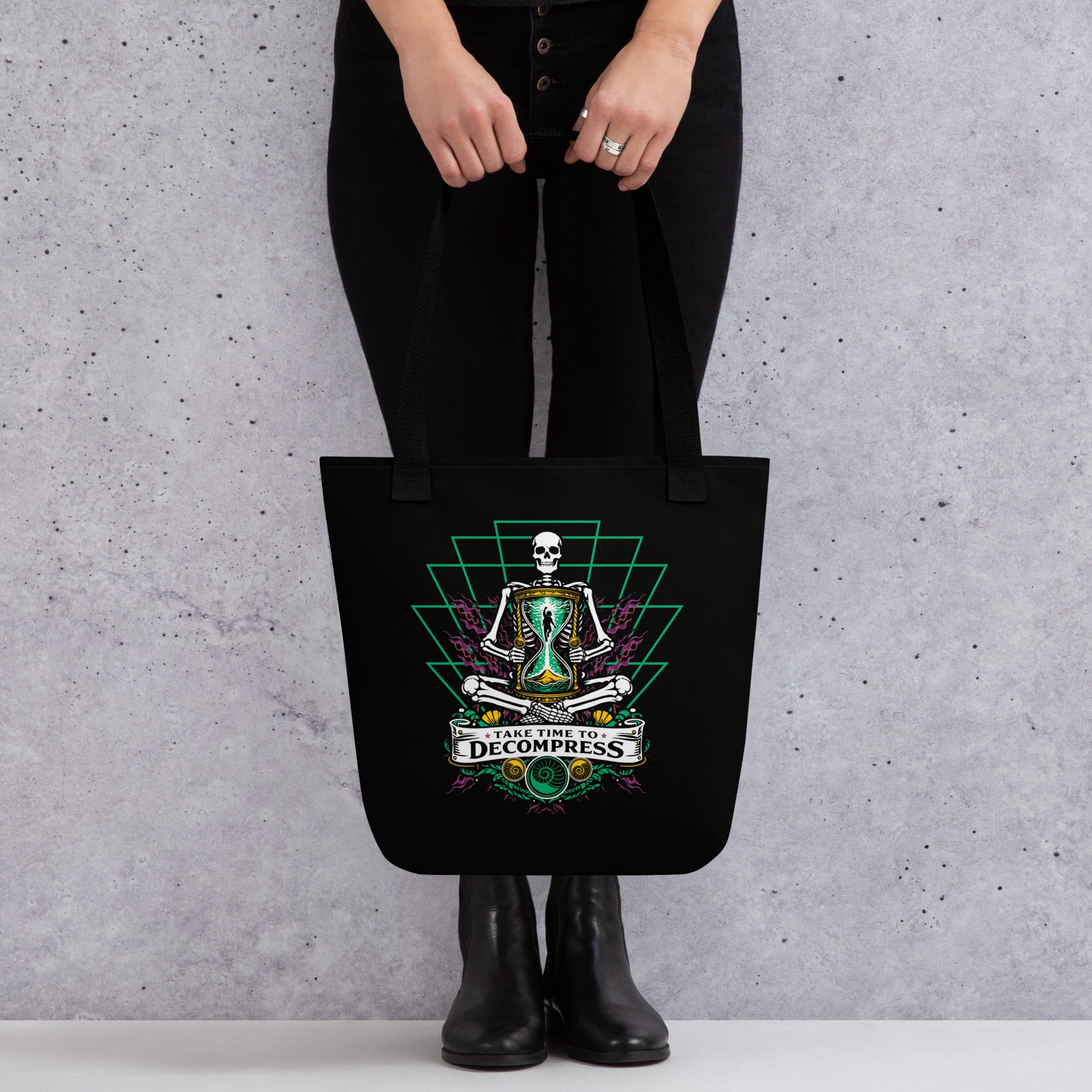 "Take Time To Decompress" black tote bag, held by model showing the back panel featuring a gold and teal "17th & Surf" hourglass logo – 17th & Surf