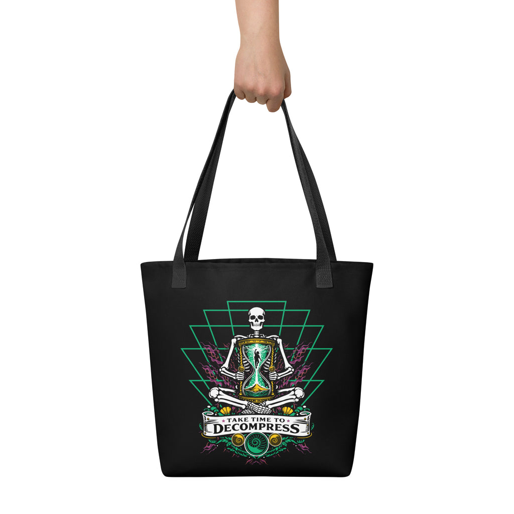 "Take Time To Decompress" black tote bag, held by model showing the front panel featuring a meditating skeleton holding a glowing hourglass with a tiny silhouette inside, framed by a green geometric pyramid pattern with purple coral accents and gold ammonite shells, above a "Take Time To Decompress" banner – 17th & Surf