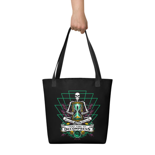 "Take Time To Decompress" black tote bag, held by model showing the front panel featuring a meditating skeleton holding a glowing hourglass with a tiny silhouette inside, framed by a green geometric pyramid pattern with purple coral accents and gold ammonite shells, above a "Take Time To Decompress" banner – 17th & Surf